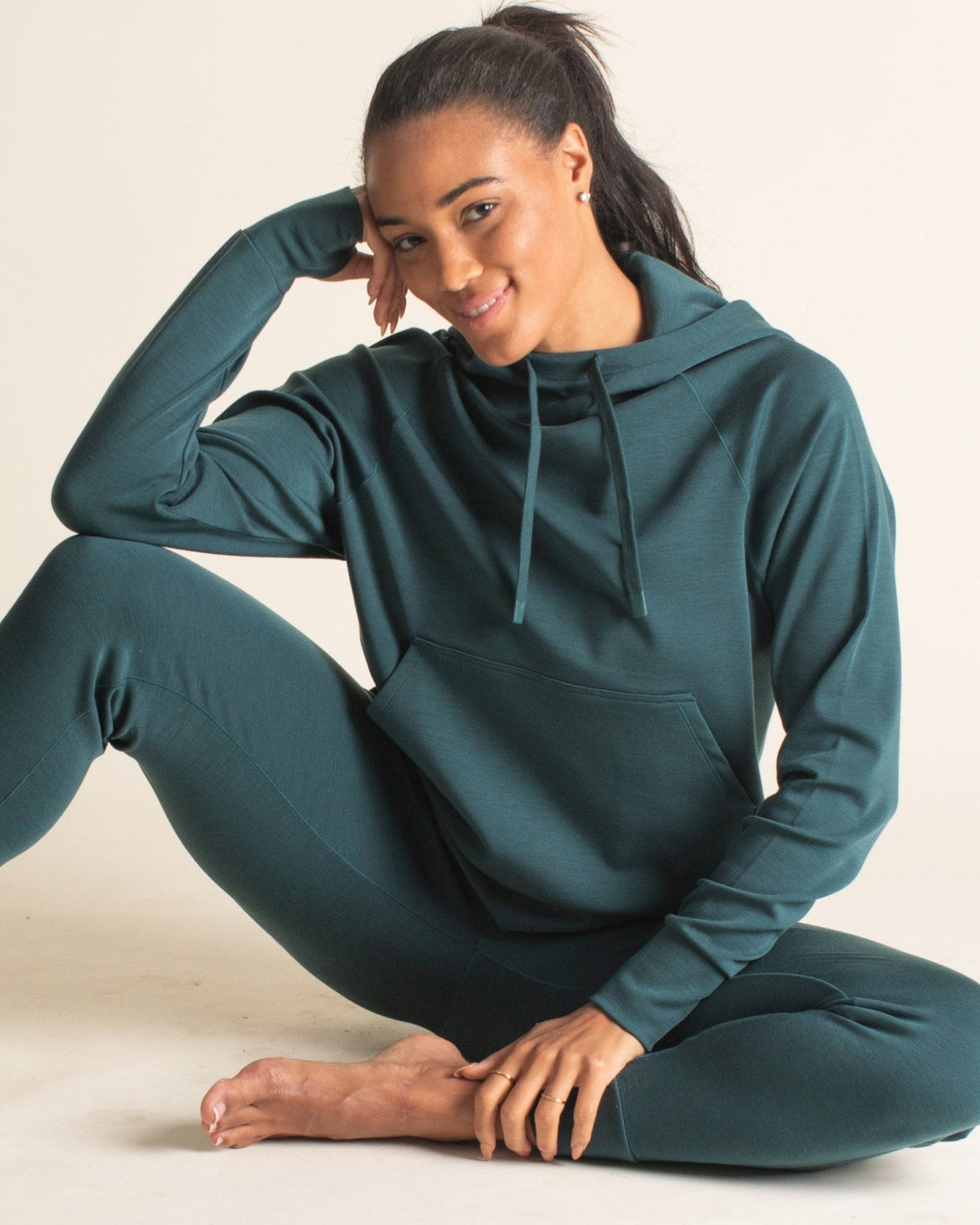 Woman sitting on the floor and smiling in Merino leggings and matching Merino wool hoodie with drawstrings, thumbholes, and a front pocket.