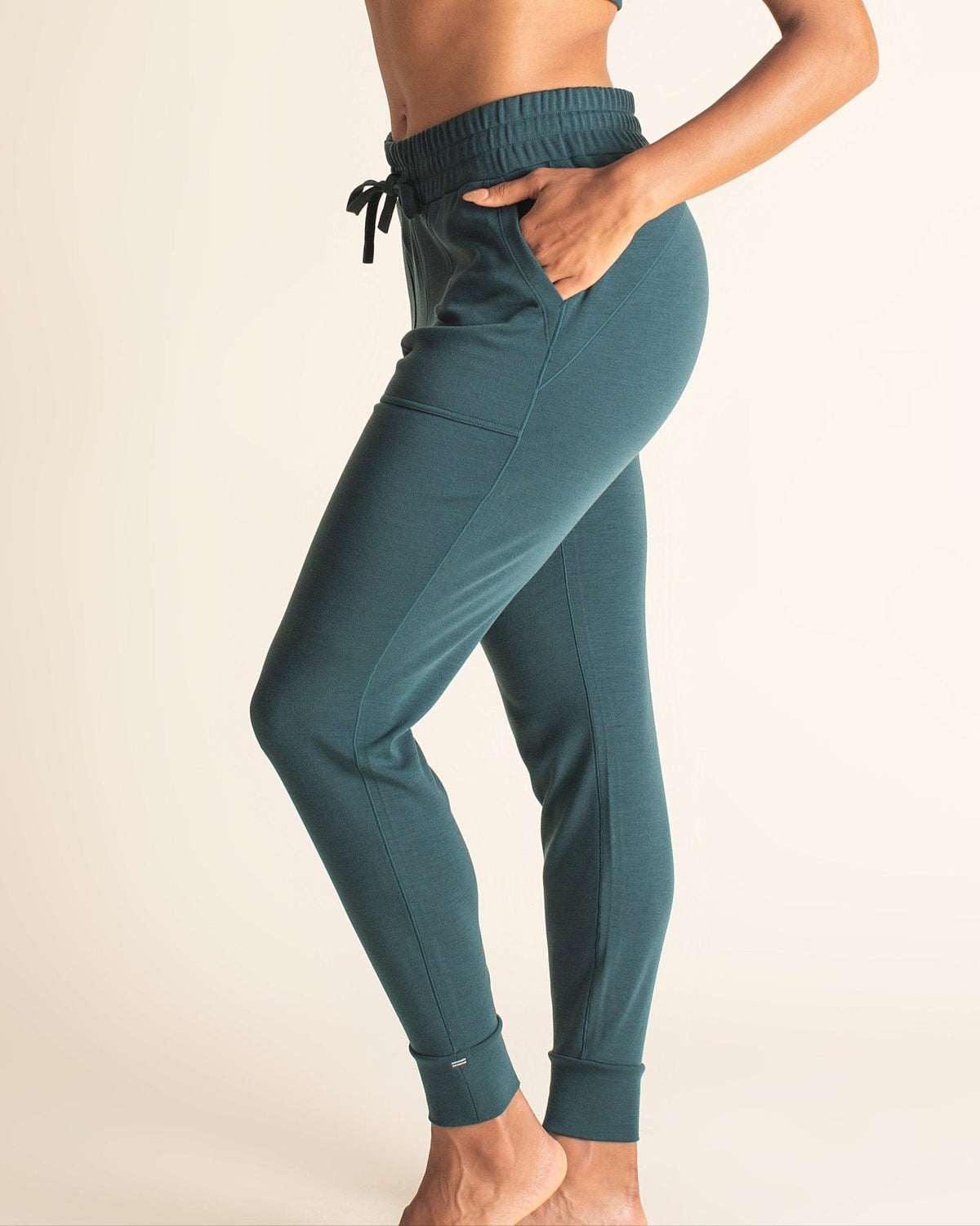 Side view of woman standing in teal BRANWYN Merino joggers with hand in pocket. Everyday wool joggers blending softness, stretch, and natural performance.