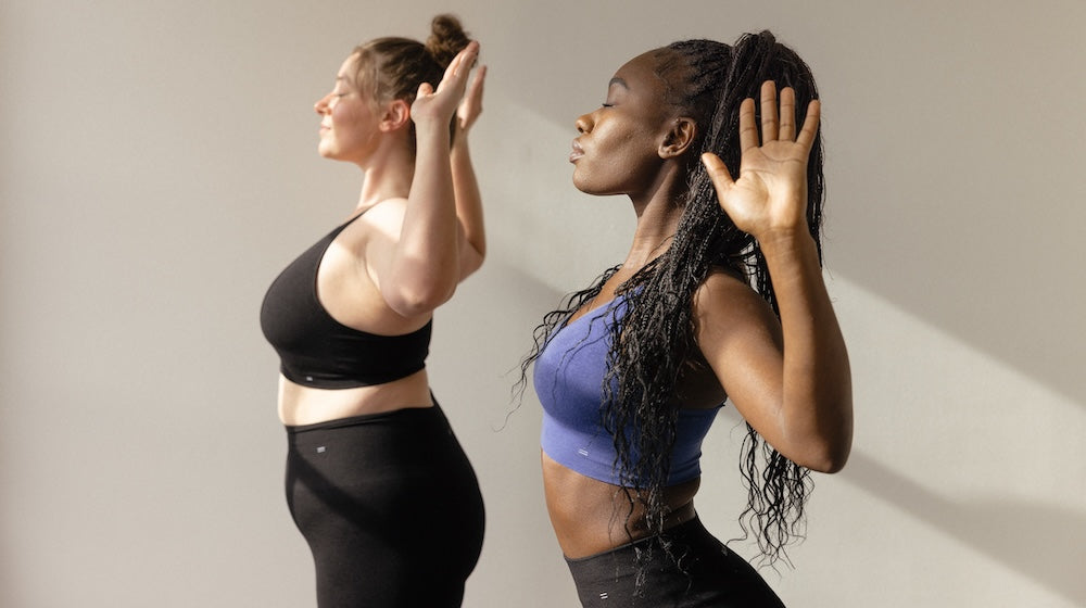 Two woman in a white room wearing leggings and bras participating in a hot yoga class.