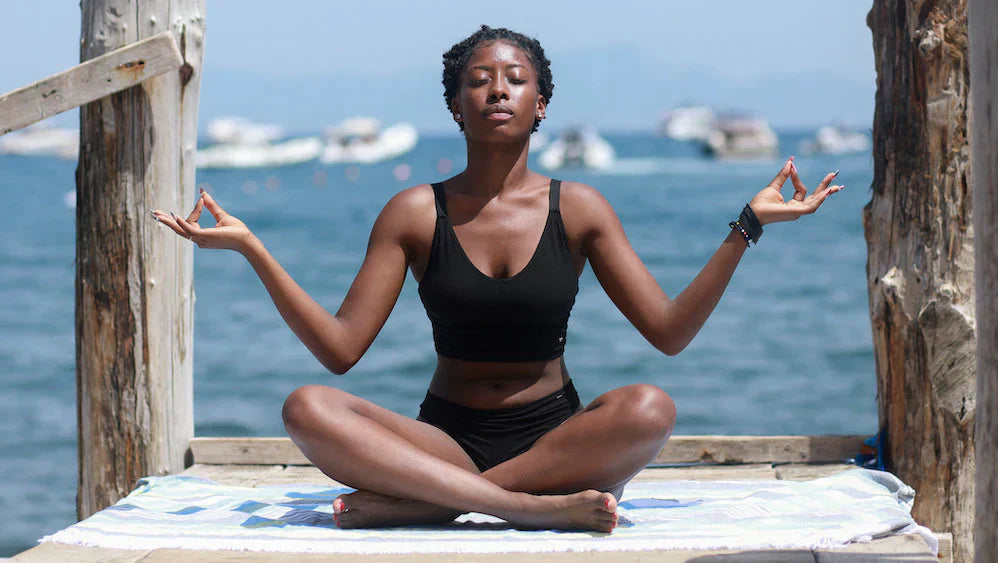 Woman meditating on a dock beside the ocean in a black Merino wool swimsuit, practicing body awareness and listening to her body's signals.