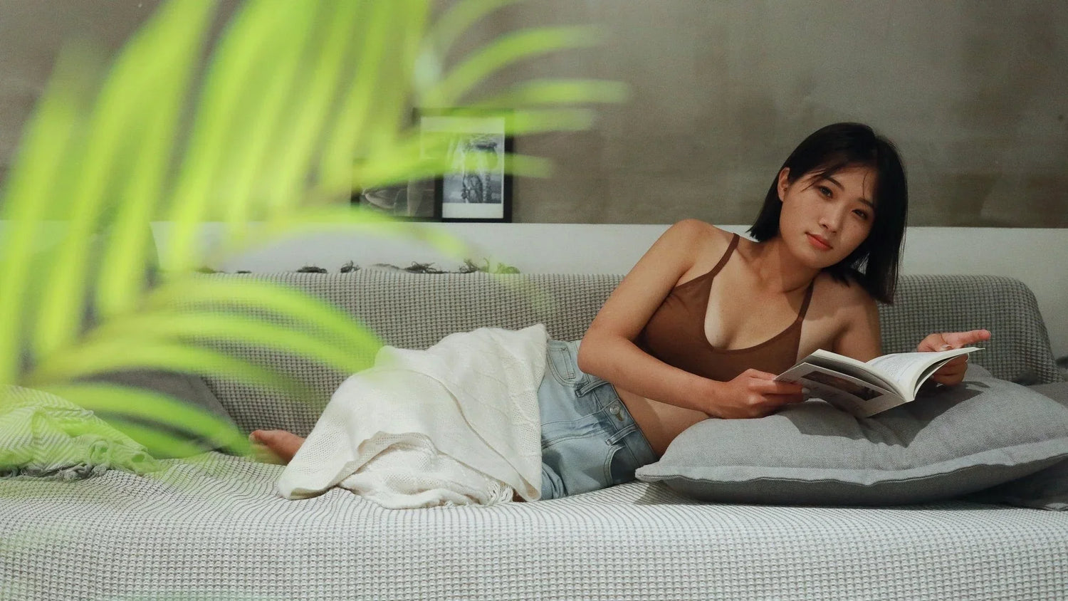 Woman laying down on a couch in shorts and a brown bra reading a book.
