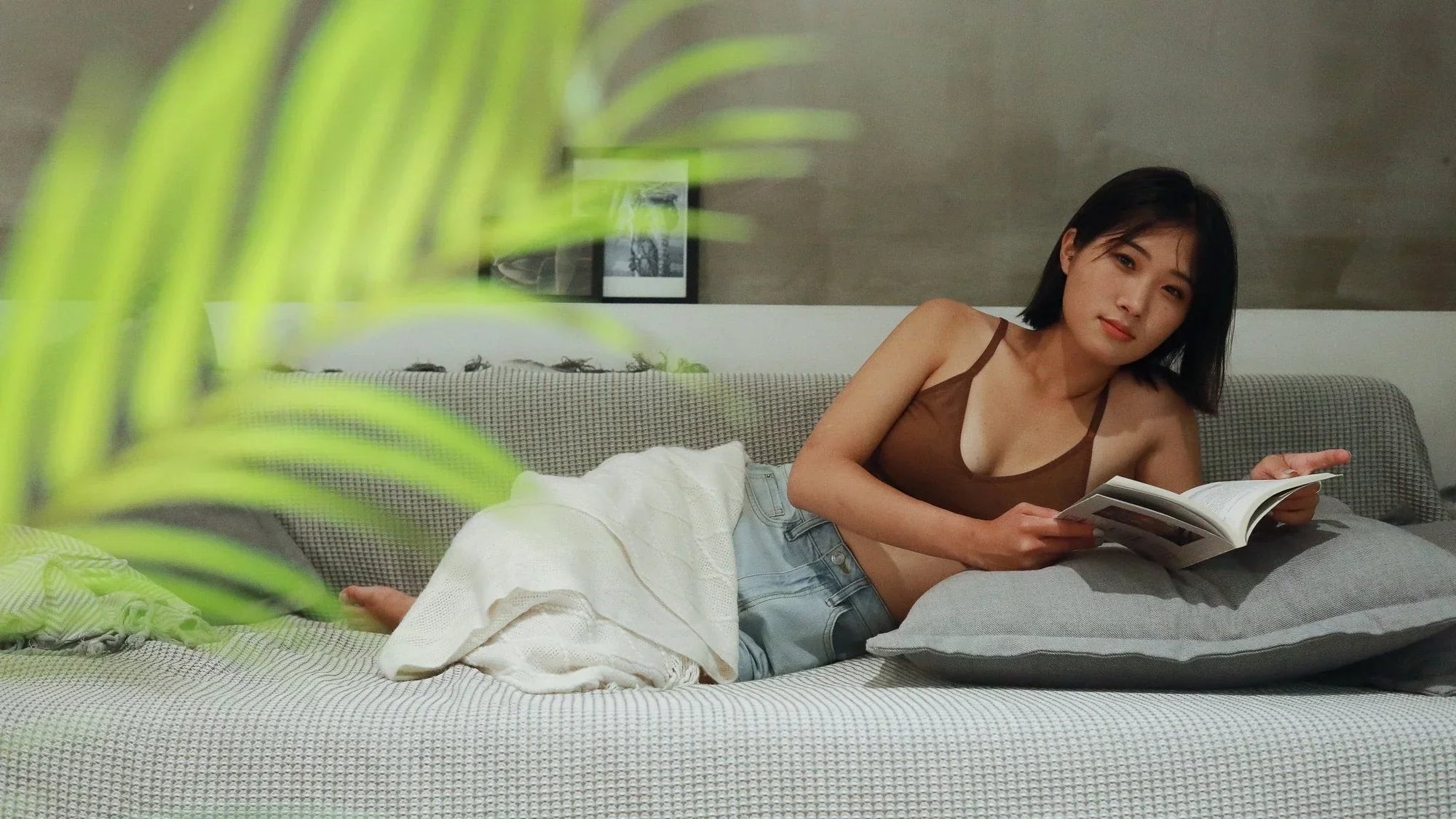 Woman laying down on a couch in shorts and a brown bra reading a book.