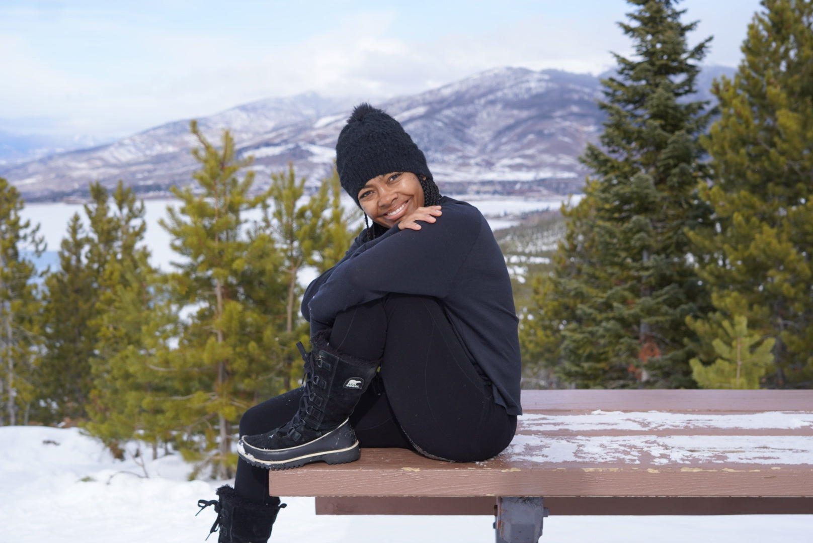 Woman in all black Merino layers, winter boots, and a beanie sitting on a picnic table in a winter mountain landscape.