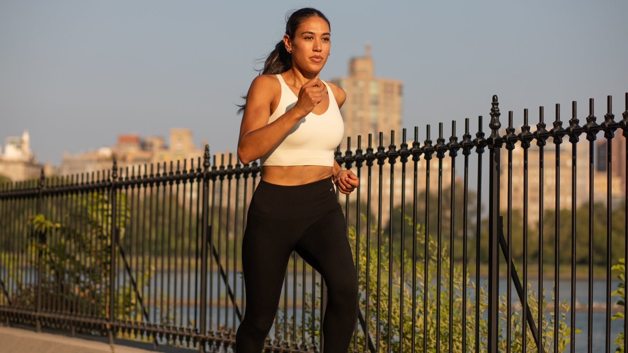 Woman running in a white Merino bra and black leggings against a city landscape. 