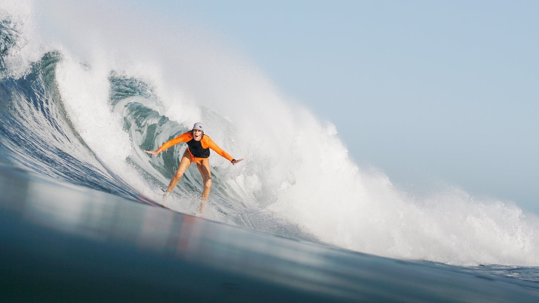 Caroline Paul in a helmet and orange suit, surfing a big wave in the ocean.