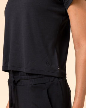 A close-up of a person wearing a black short-sleeve crop top and matching black drawstring pants against a warm beige studio background.