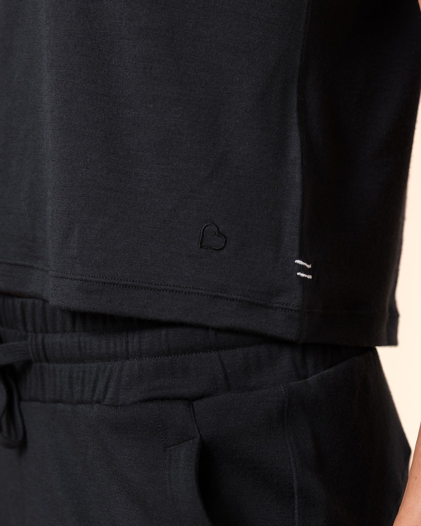 Close-up of a person wearing black lounge pants with a drawstring waistband and a black sleeveless top, against a plain warm beige background.