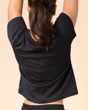 A woman wearing a black short-sleeve crop top and black wide-leg yoga pants, shown from behind in a tree pose against a solid beige studio background.