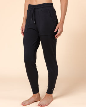 A model from the waist down wearing black wool jogger sweatpants with a drawstring waist and elastic cuffs, standing barefoot against a solid beige background.