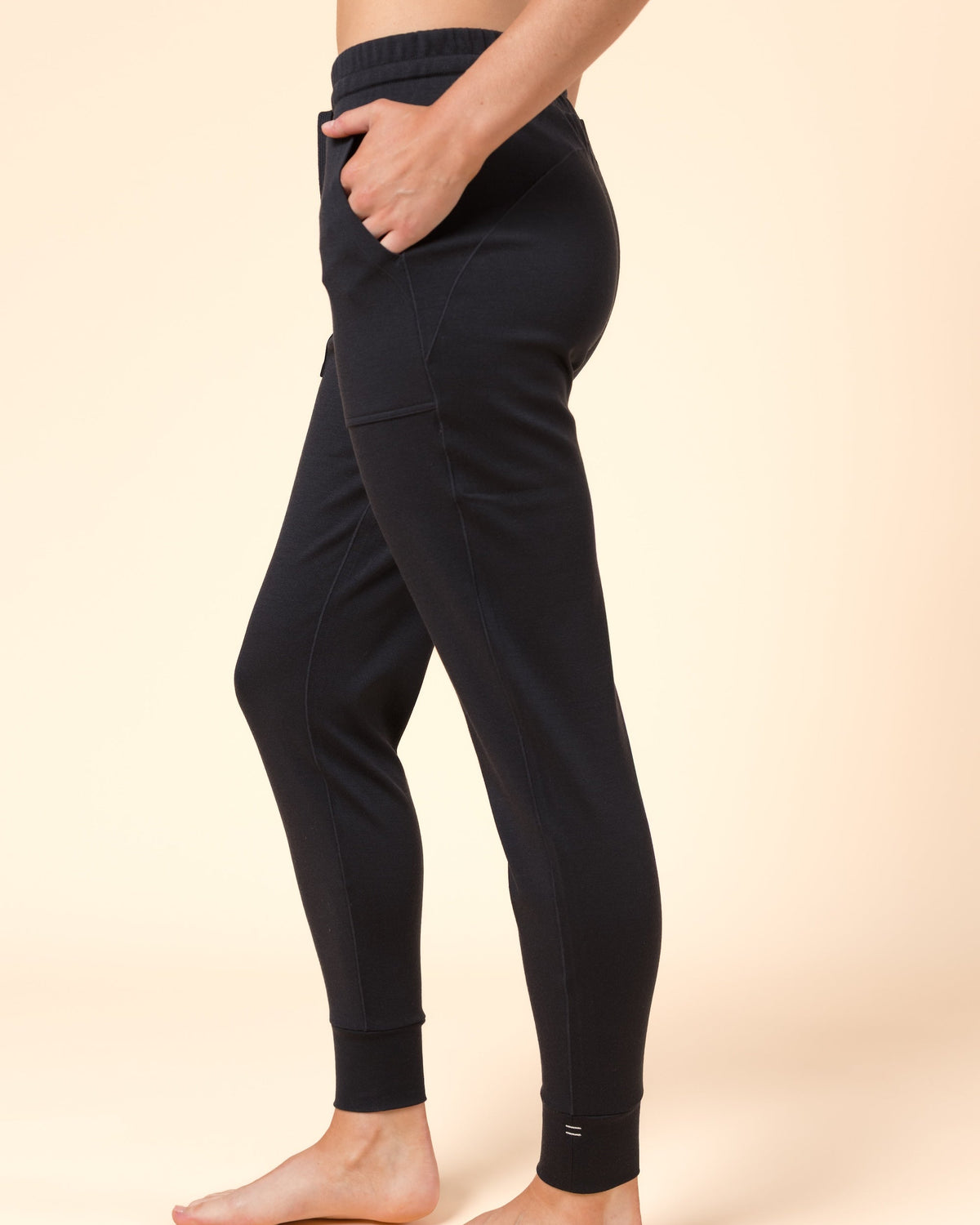 A person modeling black ankle-length pants against a warm beige studio background. The photo shows the lower torso, legs, and bare feet, with the person standing in side profile and one hand in a pocket.