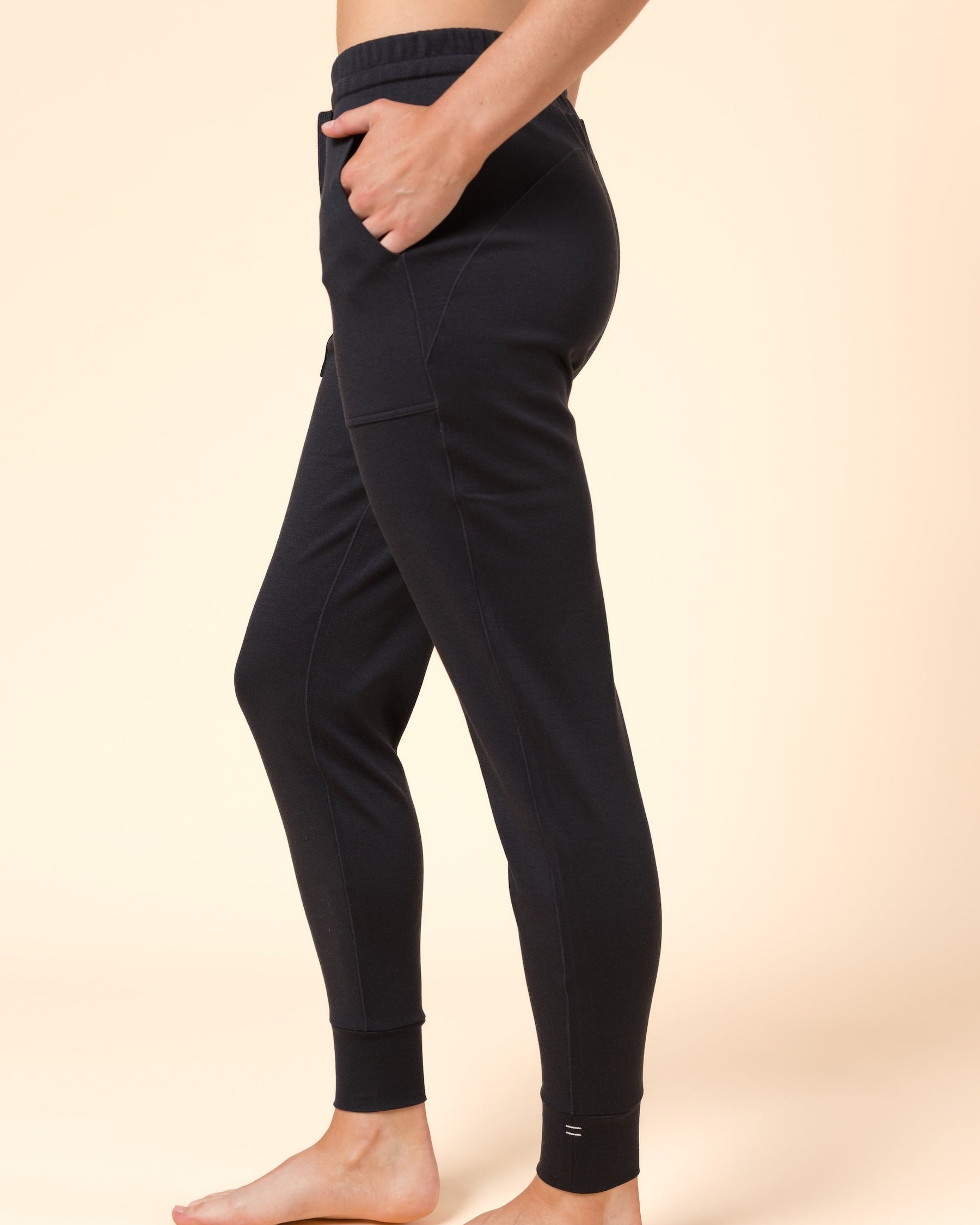 A person modeling black Merino wool joggers against a warm beige studio background. The photo shows the lower torso, legs, and bare feet, with the person standing in side profile and one hand in a pocket.
