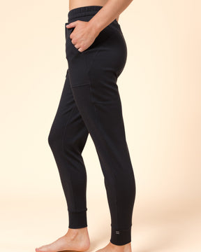 A person modeling black Merino wool joggers against a warm beige studio background. The photo shows the lower torso, legs, and bare feet, with the person standing in side profile and one hand in a pocket.