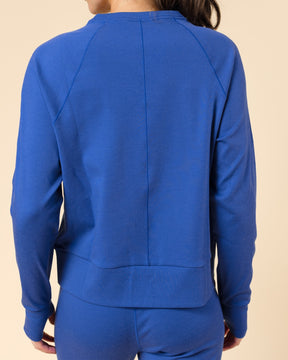A woman viewed from the back wearing a bright royal blue long-sleeve sweatshirt and matching blue pants against a plain beige studio background.