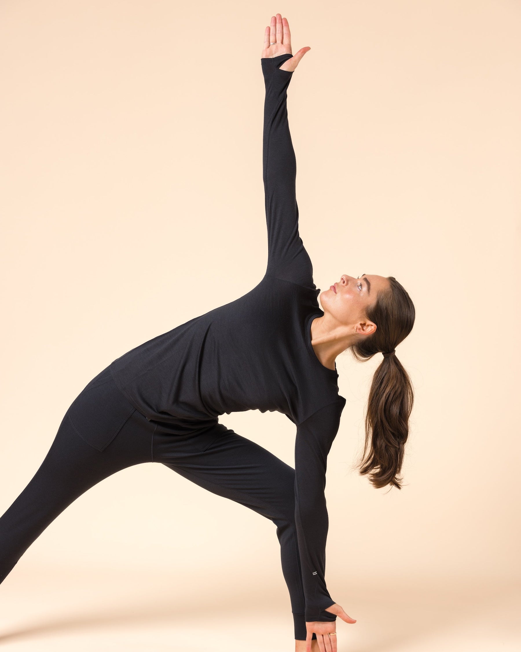 A woman in a black wool long sleeve shirt poses in a side stretch pose against a solid beige studio background.