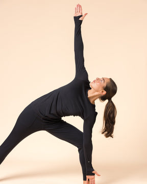 A woman in a black wool long sleeve shirt poses in a side stretch pose against a solid beige studio background.