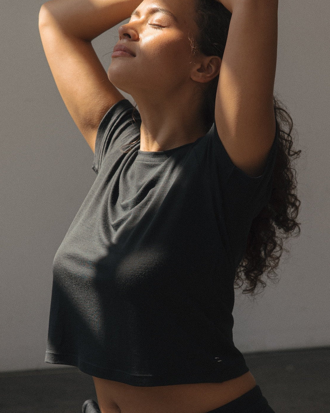 A woman in a black crop top and black leggings kneels on a yoga mat in a minimalist studio, stretching her arms overhead in a sunlit room with dramatic shadows.