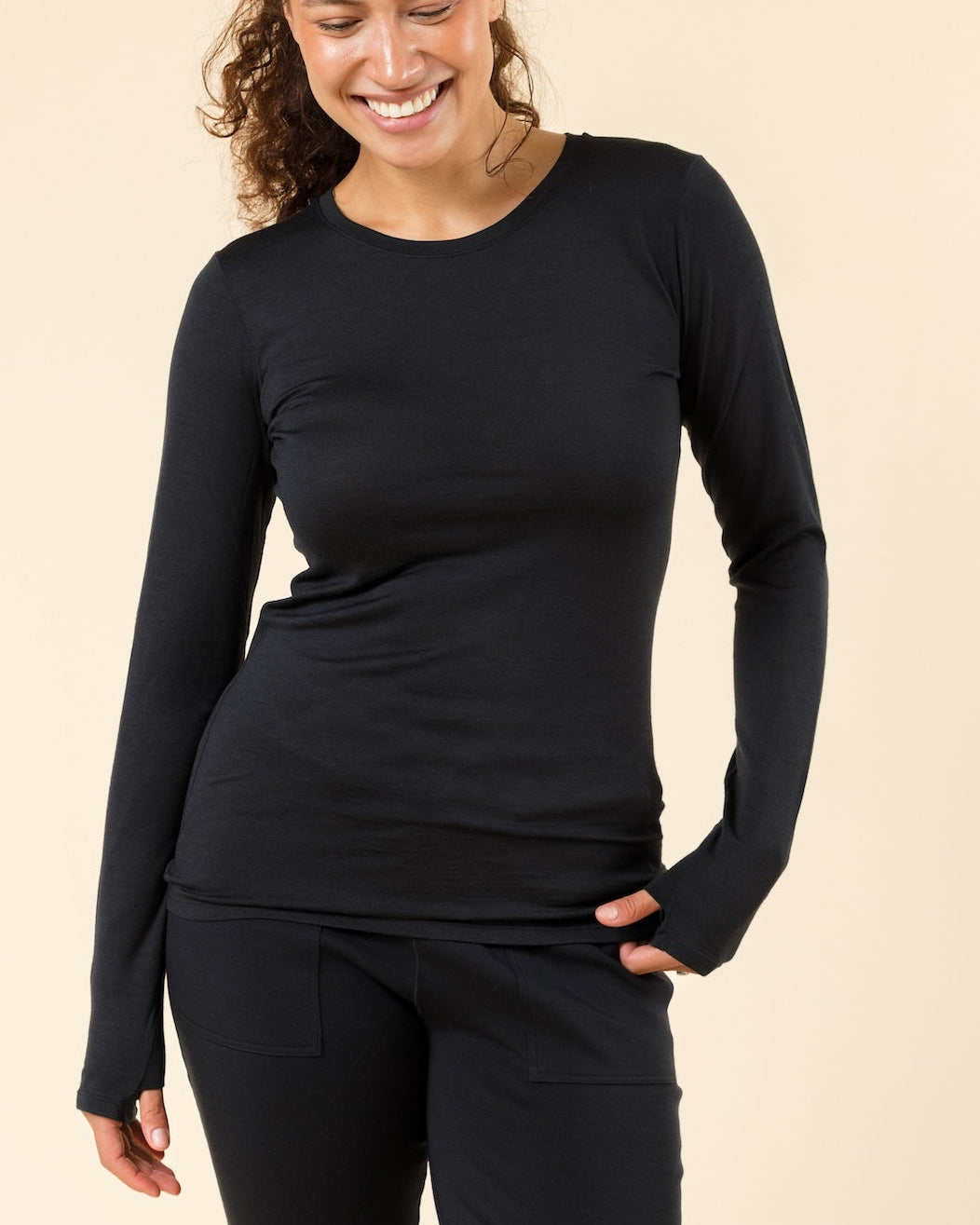 A woman smiling in a black long-sleeve crewneck merino wool baselayer against a solid beige studio background.