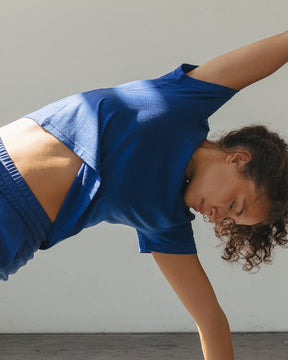 A woman in a blue activewear set performs a yoga pose on a black mat in a bright studio with large windows and strong sunlight.
