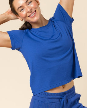 A woman posing in a matching royal blue two-piece loungewear set, featuring a short-sleeve cropped t-shirt and high-waisted jogger pants, set against a solid beige studio background.