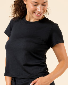 A woman wearing a black short-sleeve crewneck t-shirt and black pants, smiling and looking down against a solid beige background.