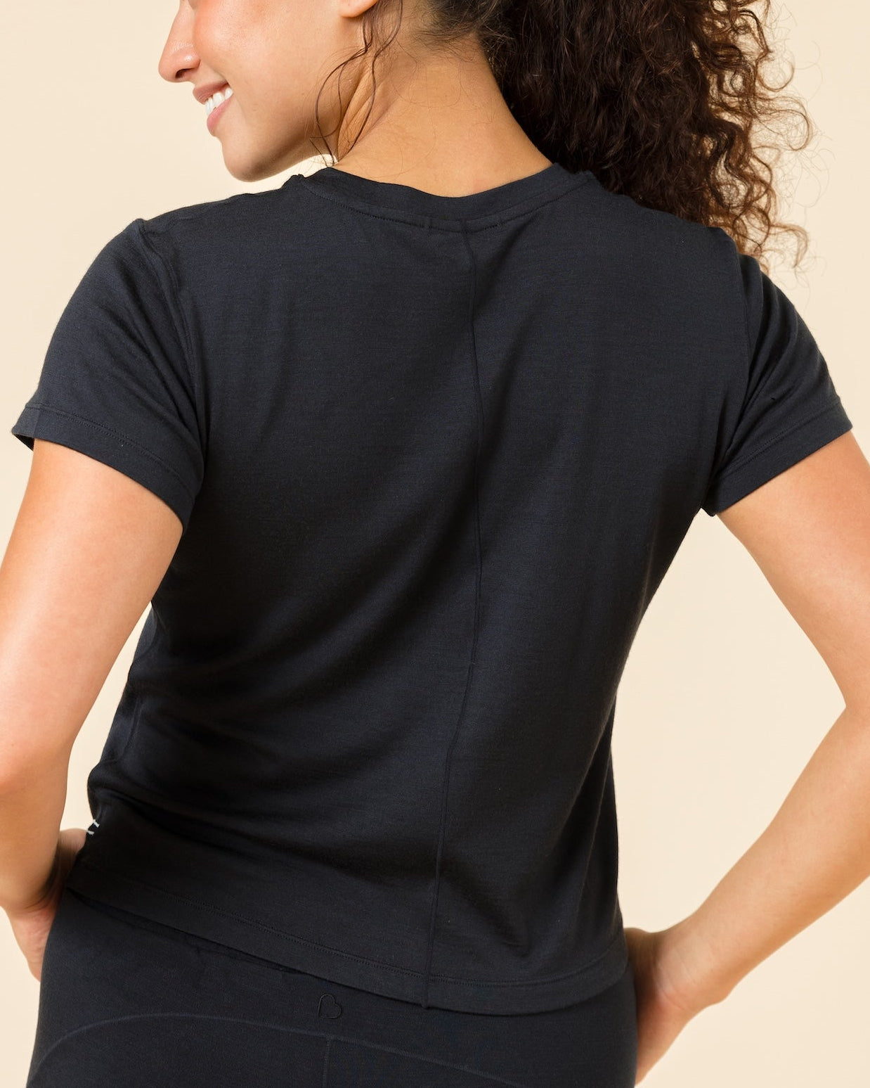 A woman with curly brown hair in a ponytail, viewed from behind, wearing a black short-sleeve athletic t-shirt and matching black leggings against a solid beige background.