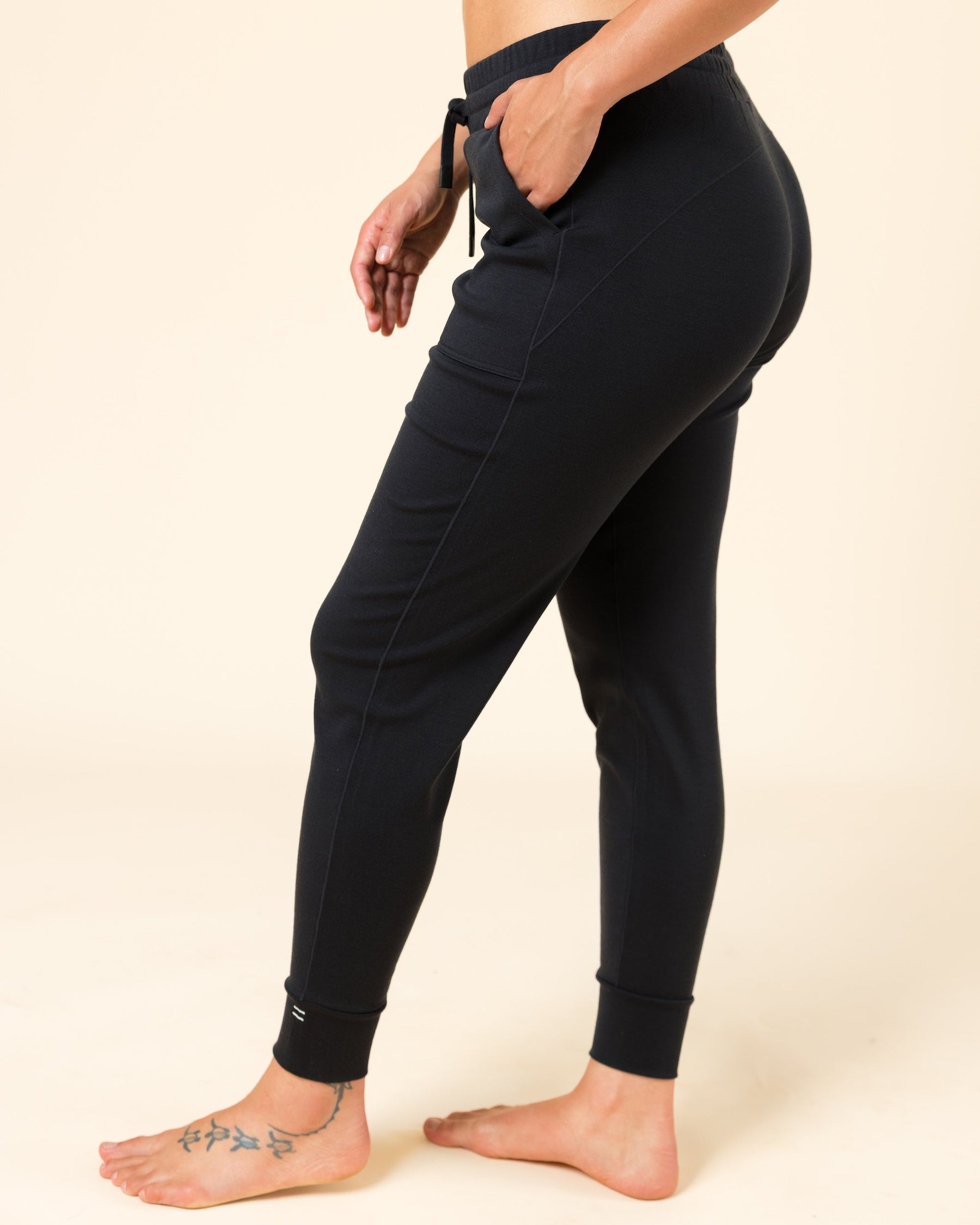 A side-profile view of a model wearing black high-waisted joggers with a drawstring waist and side pockets, standing barefoot against a solid beige background.