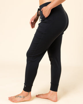 A side-profile view of a model wearing black high-waisted joggers with a drawstring waist and side pockets, standing barefoot against a solid beige background.