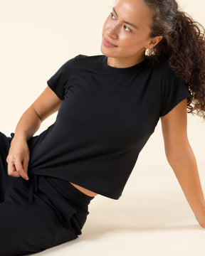 A woman with curly hair sits barefoot on a warm cream studio floor, wearing a black short-sleeve crop top and matching black wide-leg pants.