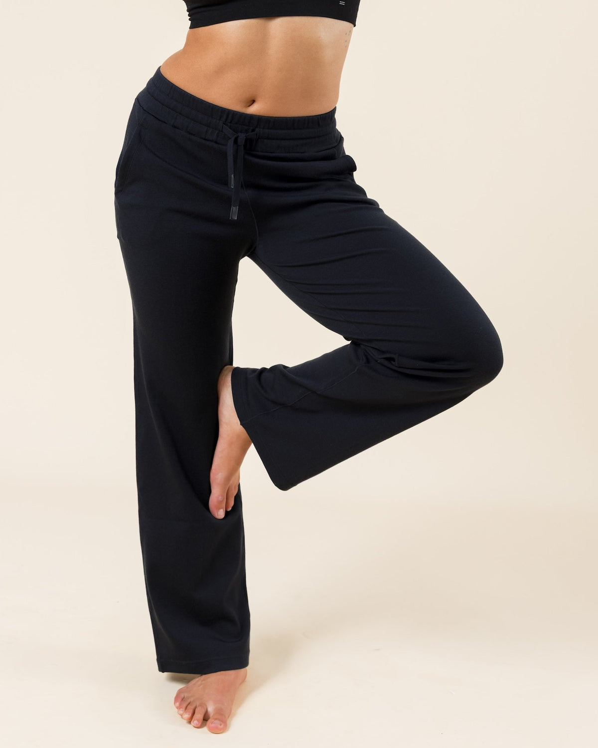 A woman modeling a black two-piece activewear set consisting of a halter crop top and matching drawstring sweatpants, standing in a yoga tree pose against a solid beige studio background.