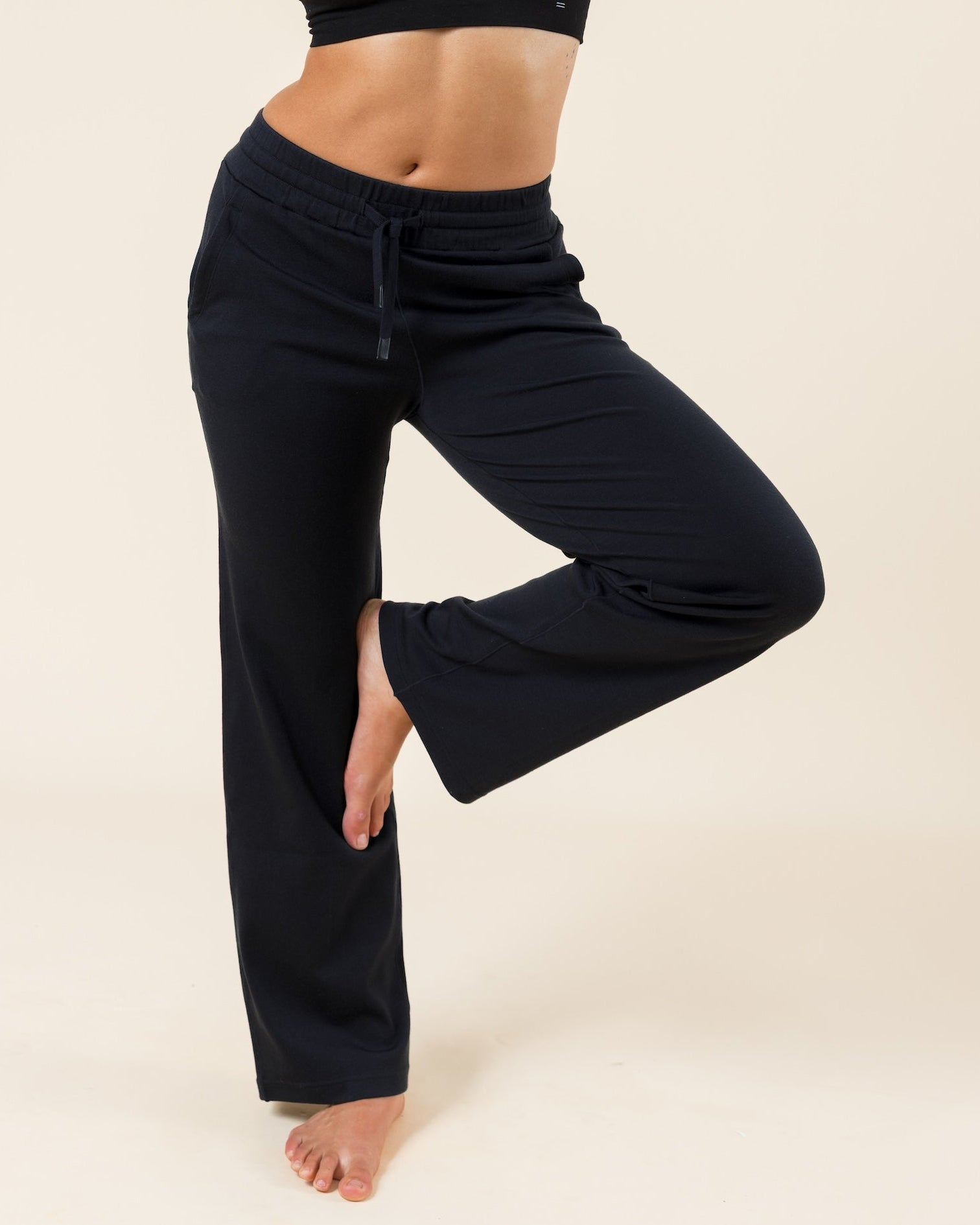 A woman modeling a black two-piece activewear set consisting of a halter crop top and matching drawstring sweatpants, standing in a yoga tree pose against a solid beige studio background.
