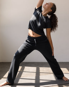 A woman in a black athletic crop top and black yoga pants poses in a side stretch yoga pose on a dark yoga mat inside a bright studio with large windows.