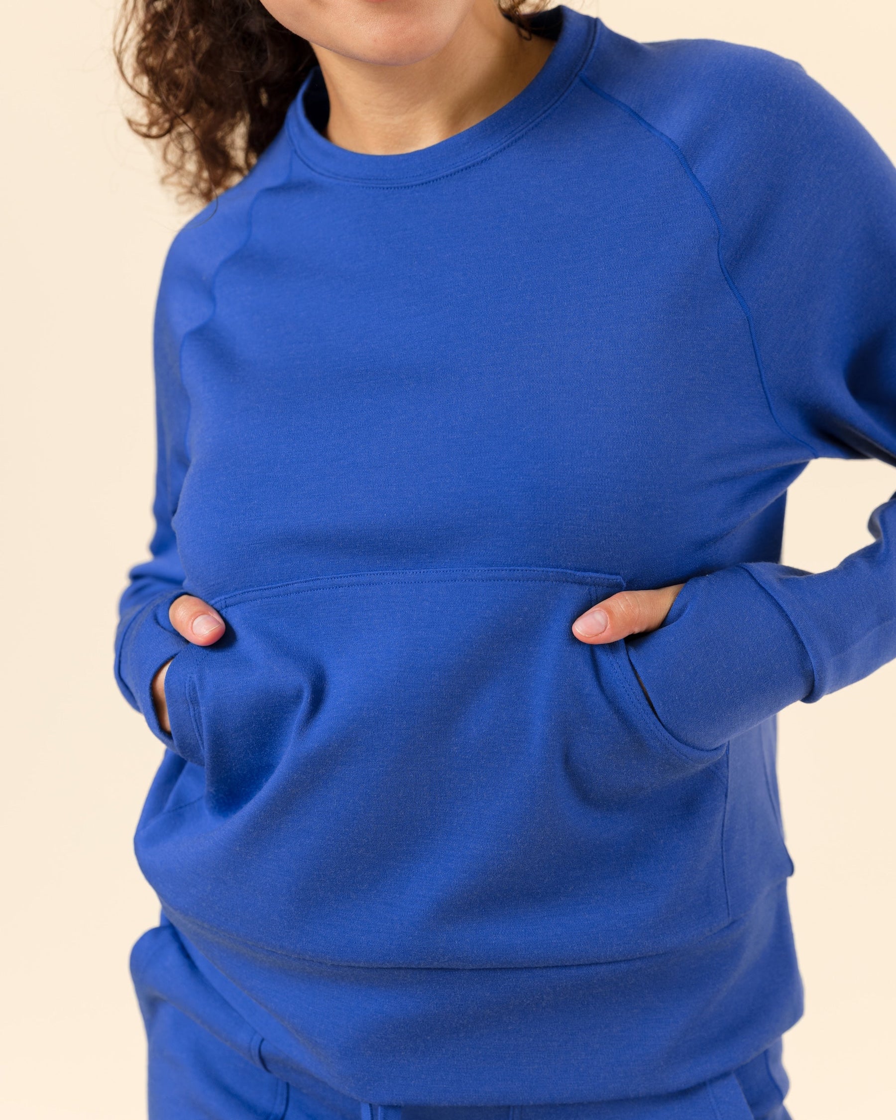 A woman wearing a bright royal blue crewneck sweatshirt with front kangaroo pockets, paired with matching blue pants, against a solid light beige background.
