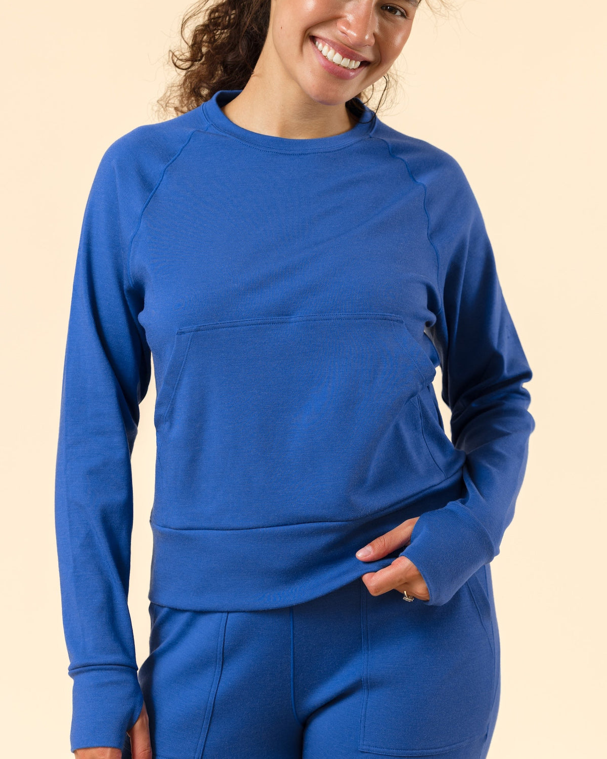 A woman modeling a matching royal blue activewear set, featuring a long-sleeve crewneck sweatshirt and matching leggings with front pockets, set against a solid light beige background.