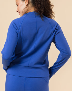 A woman with curly brown hair tied back in a ponytail, viewed from behind, wearing a matching royal blue long-sleeve top and leggings set against a solid beige background.