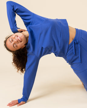 A woman in a matching royal blue activewear set, consisting of a long-sleeve crop top and high-waisted leggings, performing a side stretch pose on a solid beige studio background.