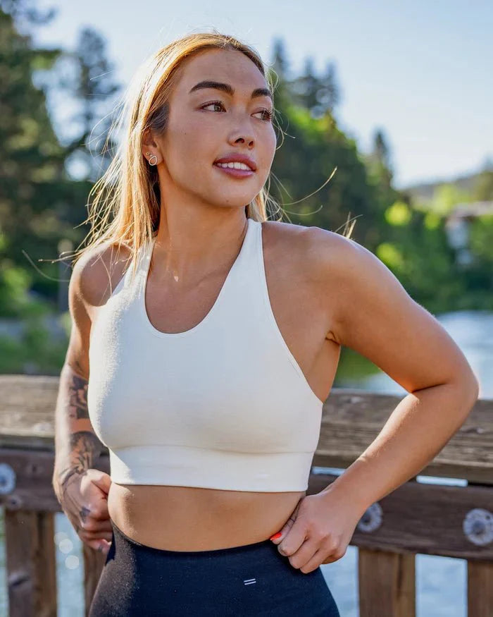 Woman standing beside a river in BRANWYN Racerback Sports Bra in natural white. Merino seamless sports bra with sleeve for pads.