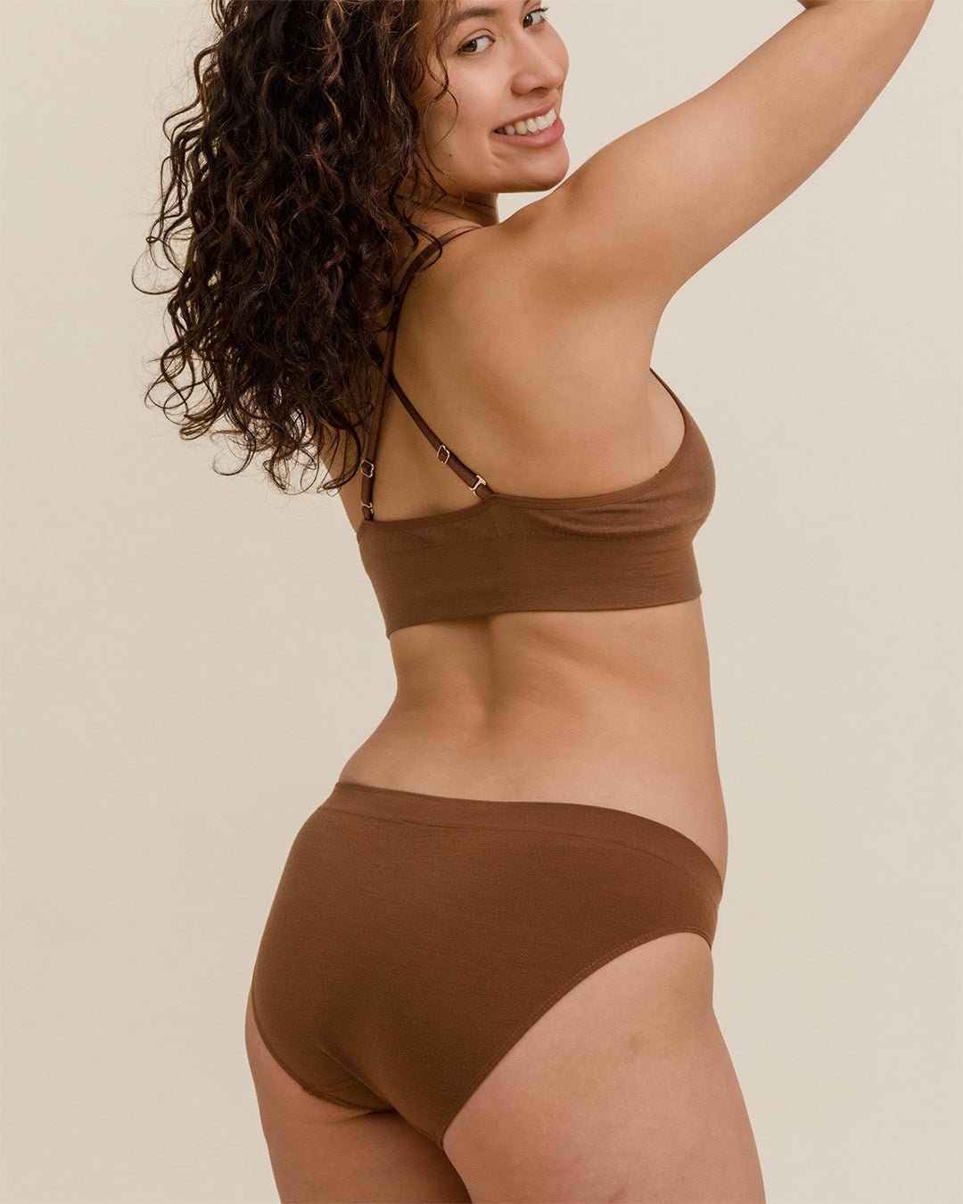 Back view of woman with curly hair smiling in BRANWYN seamless Merino wool bra and underwear set in dark nude brown
