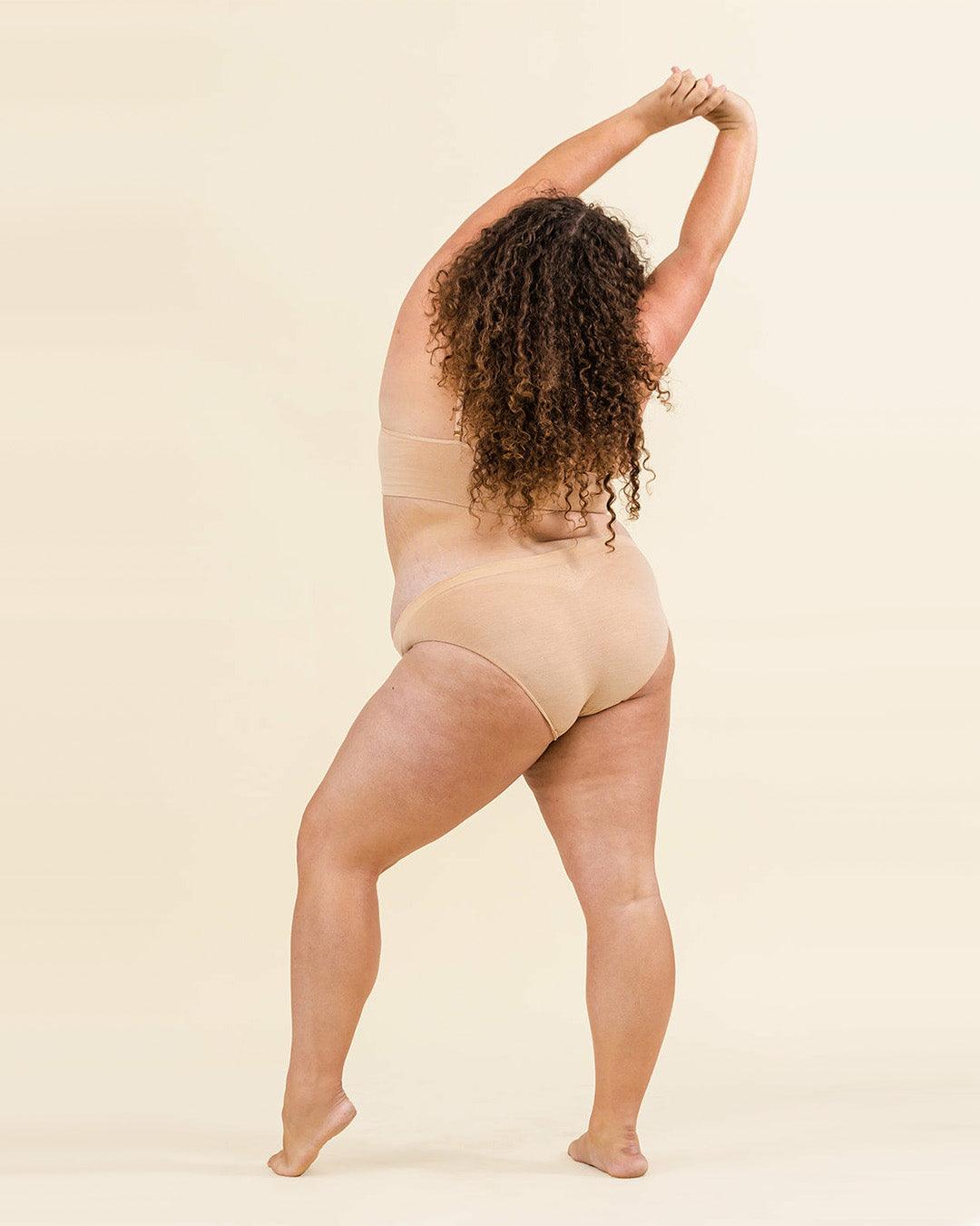 Woman posing with arms raised overhead in BRANWYN women’s seamless Merino wool bikini underwear set in light nude.