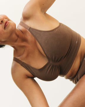 A woman in a brown supportive bralette and matching shorts underwear performing a side stretch against a plain off-white background.