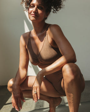 A woman with curly hair crouches on a gray yoga mat in a sunlit room, wearing a brown padded bralette and matching underwear.