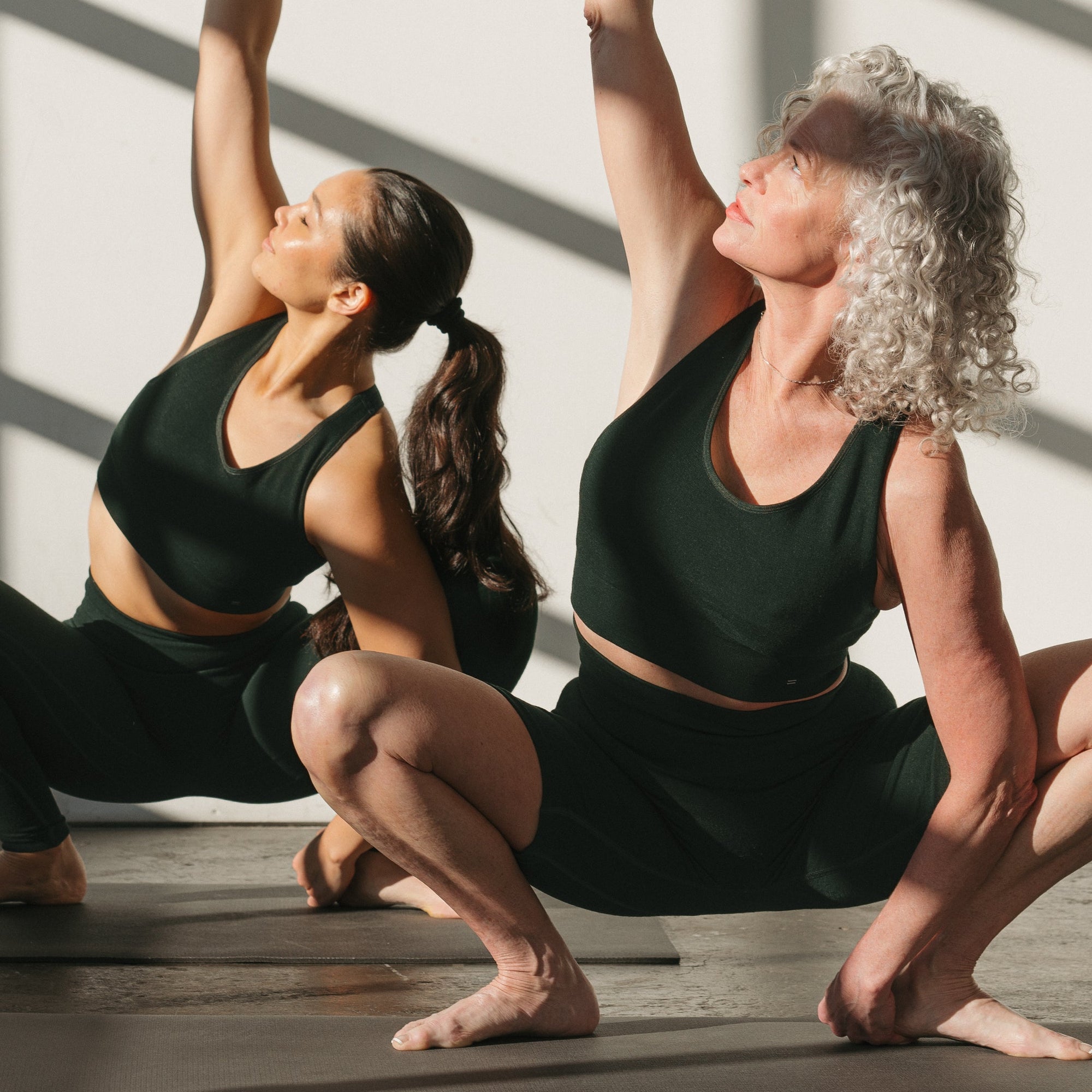 Two women in green athletic wear performing a yoga pose in a well-lit room with large windows.