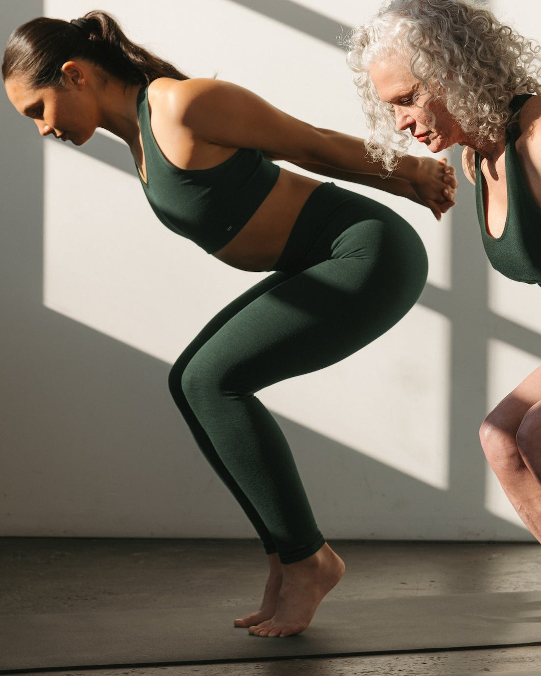 Two women stretching together in BRANWYN seamless Merino wool Racerback Sports Bras and Compressive Leggings in Deep Willow green