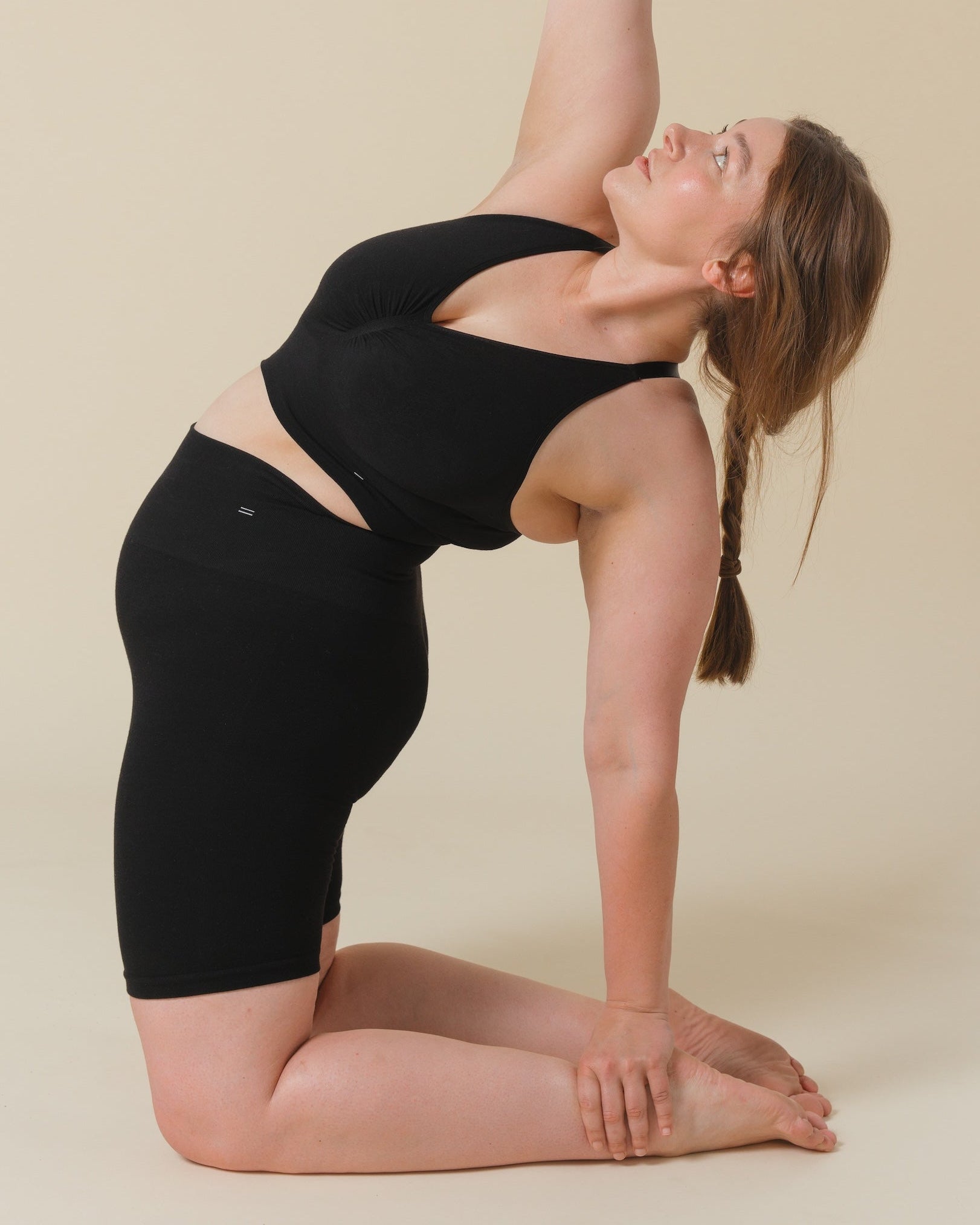 A woman in a black Merino wool padded bralette and matching black shorts performs a yoga backbend pose against a solid beige studio background.