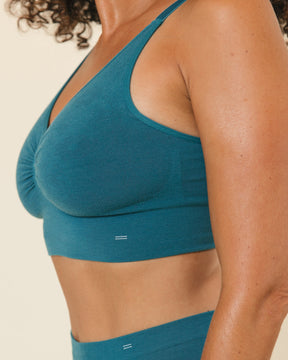 Side profile view of a woman wearing the BRANWYN Transformative Teal Supreme Bra -a pull-over seamless Merino wool with BRANWYN logo detail, against a cream background.