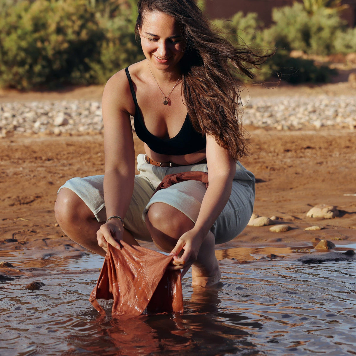 A woman crouches in shallow water, washing her Merino wool sports bra in a river. 