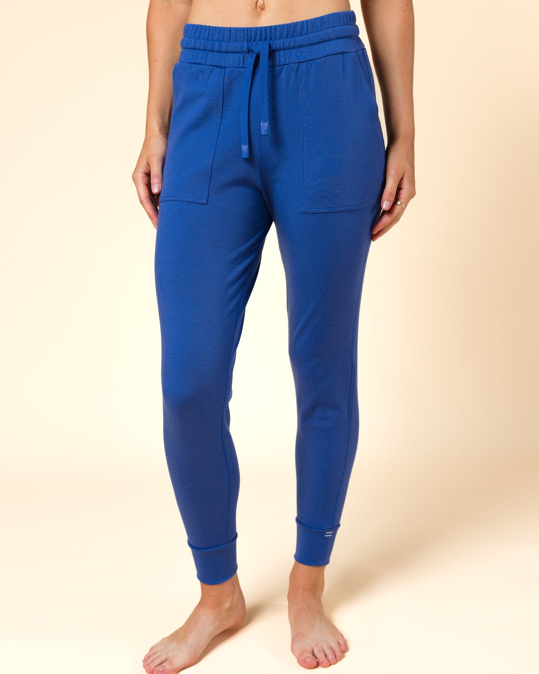A person modeling a pair of bright blue wool joggers with an elastic waistband, drawstring, side pockets, and ribbed ankle cuffs, standing barefoot against a plain beige studio background.