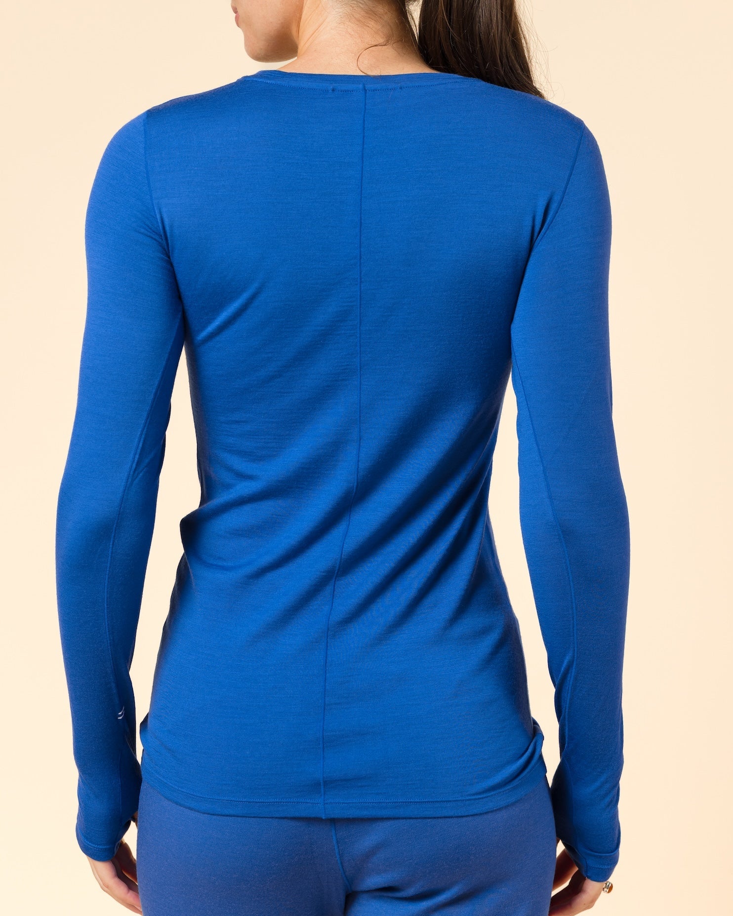 A woman viewed from the back wearing a fitted royal blue long-sleeve merino wool baselayer top and matching blue leggings against a plain warm beige studio background.