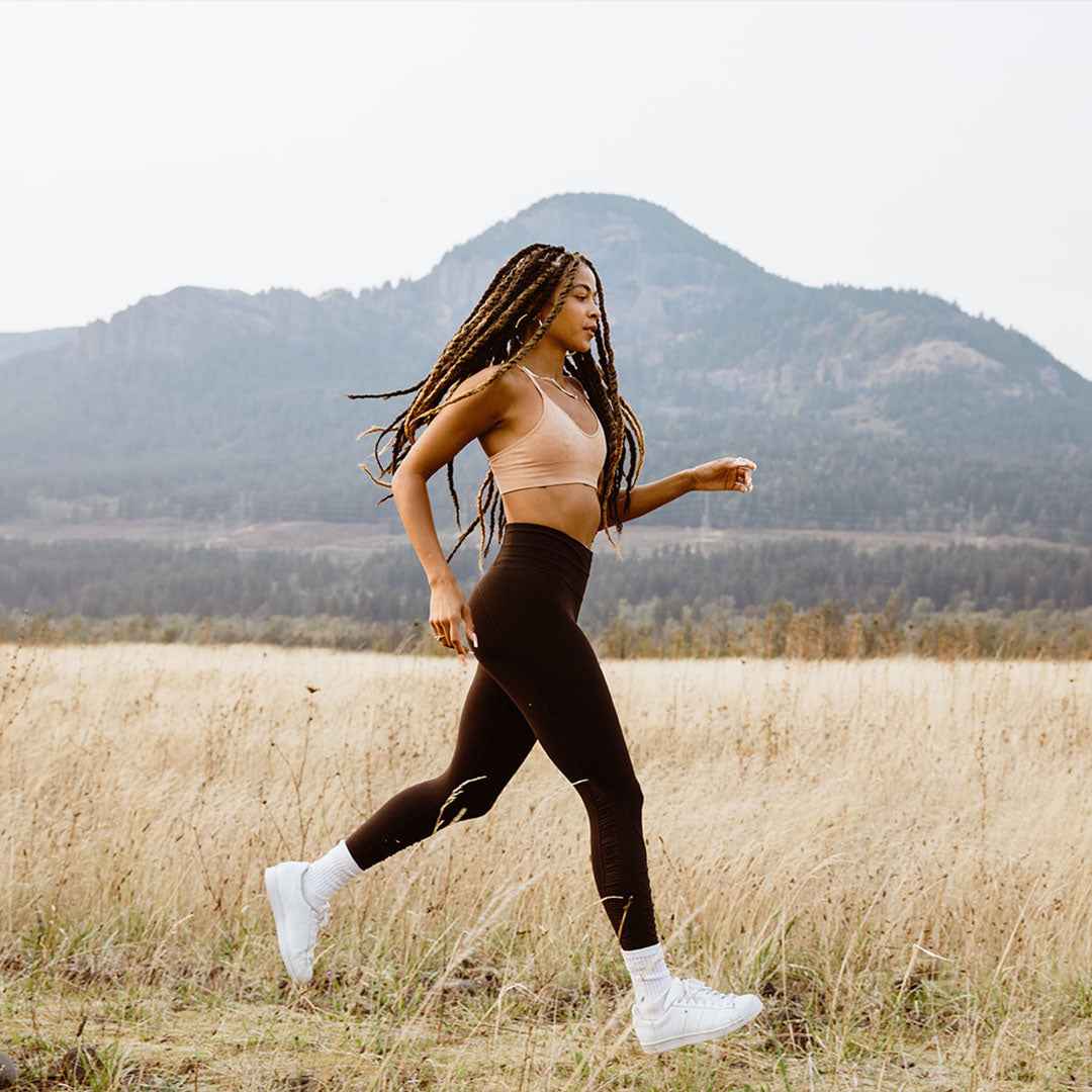 Woman with long braids running in a field wearing a nude Merino bralette, brown leggings and white sneakers.