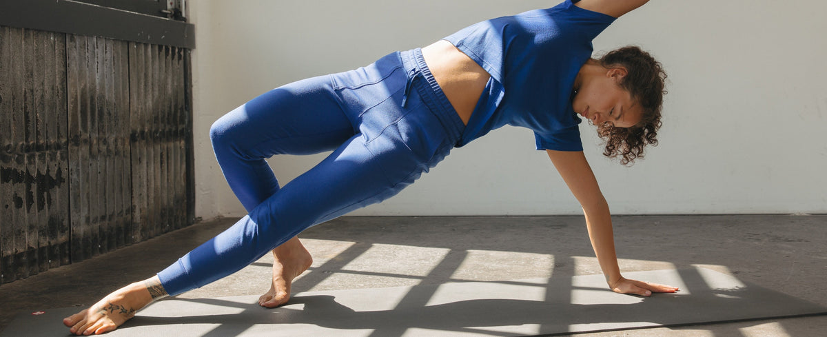 A woman in a BRANWYN's Merino wool boxy tee and joggers performs a yoga pose on a black mat in a bright studio with large windows and strong sunlight.